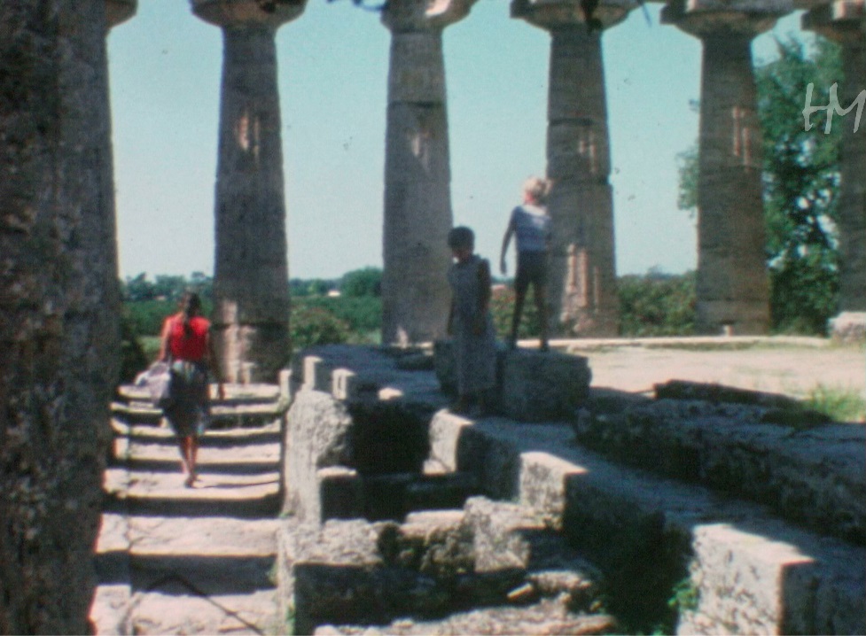 Grazia di spalle mentre i bambini giocano disordinatamente sulle rovine di Paestum. Giuseppe Lenzi, HMLENZGIU-0046, 8mm, 1972-1980, conservato presso Home Movies &ndash; Archivio Nazionale del Film di Famiglia.
